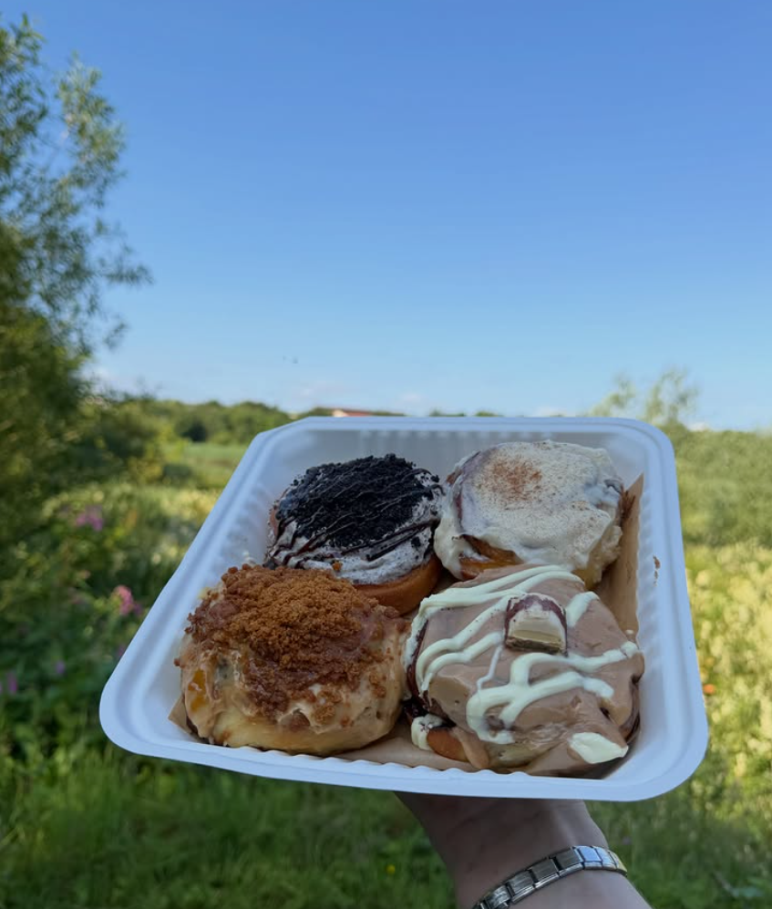 A tray of four freshly iced cinnamon rolls — Oreo, Classic, Biscoff and Kinder Bueno — held up in front of the forest